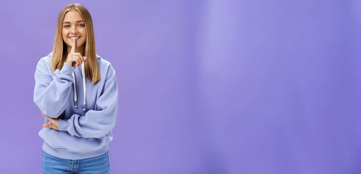 Portrait of joyful cute feminine young woman in hoodie smiling happily showing shush gesture hiding surprise asking keep secret standing amused and carefree against purple background. Copy space