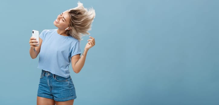 Lifestyle. Girl enjoying cool bits in brand new wireless earphones advertising earbuds in own blog recodring video via smartphone dancing from joy and delight smiling listening music over blue background.