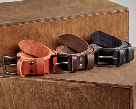 Closeup of collection of three rolled leather belts of different colors with metal buckles and DAD embossing on loops, displayed on wooden riser
