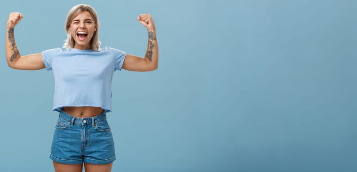 Strong women stand up and fight for rights. Portrait of happy entertained and cool young female athletic blonde with tattoos winking and smiling showing muscles and biceps over blue wall.