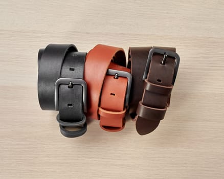 Three folded leather belts of different colors with stylish buckles and DAD embossing on loop arranged on light wooden background. Stylish personalized handcrafted mens accessories