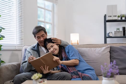 Asian senior couple reading book diary together, sitting on cozy sofa in home interior. Happy retirement activity lifestyle at home.