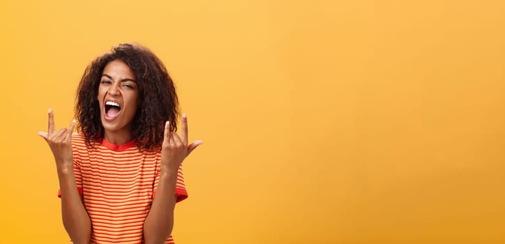 Waist-up shot of amazed happy stylish african american woman feeling awesome rocking on party yelling from joy and satisfaction showing rock n roll gesture posing over orange background. Copy space