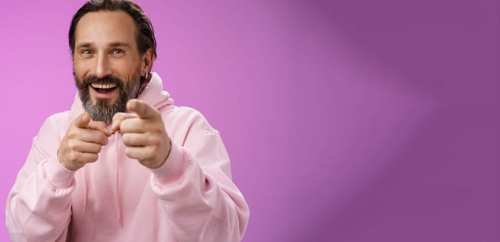 Funny amused carefree happy adult handsome man pranking friend fool around pointing camera index fingers greeting choosing you laughing joyfully having fu, standing purple background rejoicing.