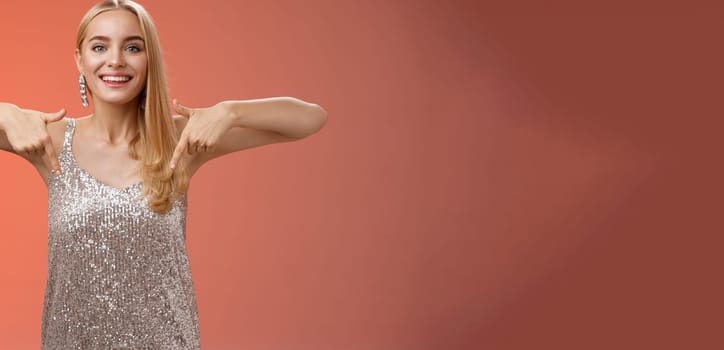 Lifestyle. Attractive glamour blond woman in silver glittering dress pointing down smiling excited showing awesome party place hang-out inviting try-out standing pleased red background enjoying perfect evening.