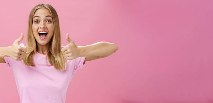 Supportive girlfriend cheering for favorite player showing thumbs up with happy positive smile giving approval and showing she likes perfect idea posing amused and enthusiastic against pink background. Lifestyle.