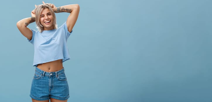 Lifestyle. Studio shot of cute attractive and joyful stylish blond woman with tattoos on arms winking and smiling broadly holding index fingers on head making horns standing in summer outfit over blue wall.