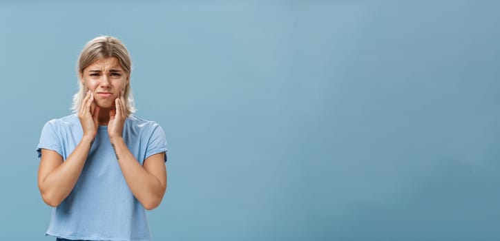 Girl feeling pain in teeth after drinking hot coffee. Displeased unhappy troubled young girl with blond hair frowning feeling discomfort in mouth holding hands on cheeks suffering toothache.