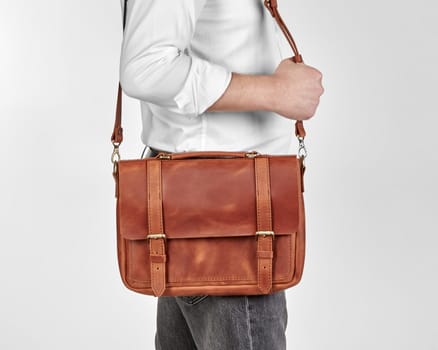 Man in white shirt and jeans carrying comfortable copper-colored leather messenger bag over shoulder with embossed initials JEN. Classic stylish artisanal accessory for business man