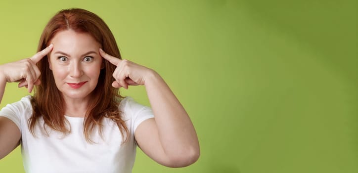 Woman playfully staring funny control your mind. Silly redhead middle-aged female touch temples popping eyes smiling delighted read thoughts trying guess intrigued what thinking green background.