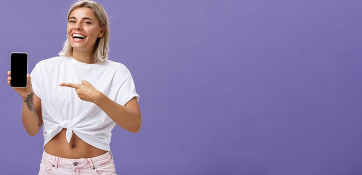 Satisfied happy beautiful tanned woman with blond hairstyle and tattoos holding smartphone showing device screen and pointing at gadget with index finger smiling at camera over purple wall. Technology and advertising concept