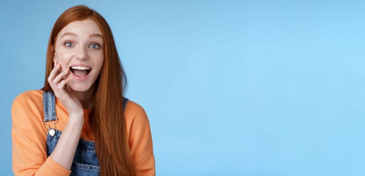 Excited enthusiasitc charismatic redhead female friend look surprised amused touch cheek open mouth thrilled discussing future prom rejoicing standing blue background joyful delighted.