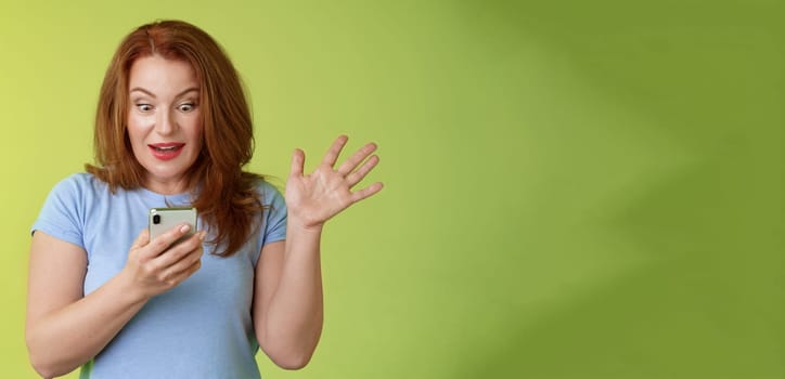 Surprised enthusiastic happy redhead mature middle-aged woman. receive excellent message read perfect news social media hold smartphone stare astonished pleased mobile phone screen raise hand triumph.