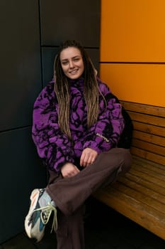 Stylish young woman with dreadlocks stands against the background of an orange house.