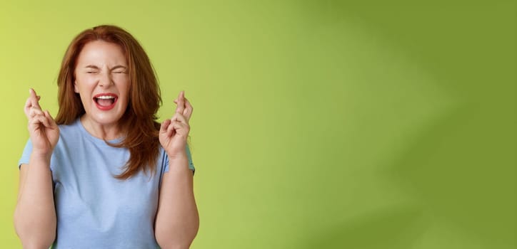 Woman wants win badly. Enthusiastic lucky redhead middle-aged 50s female pleading implore god make dream come true cross fingers good luck wishing closed eyes open mouth excitement green background.