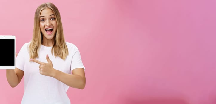Woman giving recommendation buy digital tablet pointing at device screen with index finger smiling enthusiastic and pleased telling own opinion about gadget perfomance posing delighted over pink wall. Lifestyle.