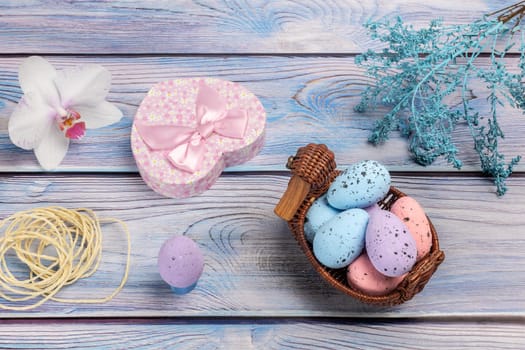 Colorful Easter eggs in a wicker basket, a gift box, an orchid flower, a rope and decoration plants on boards. Top view.