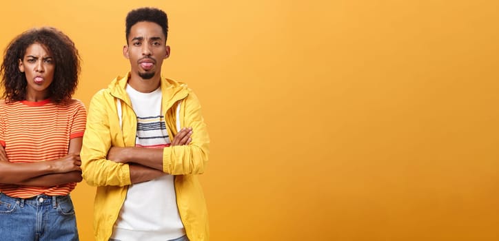 Indoor shot of african american siblings being displeased and annoyed showing bad tempber behaving childish sticking out tongue standing together with crossed hands on chest over orange background. Lifestyle.