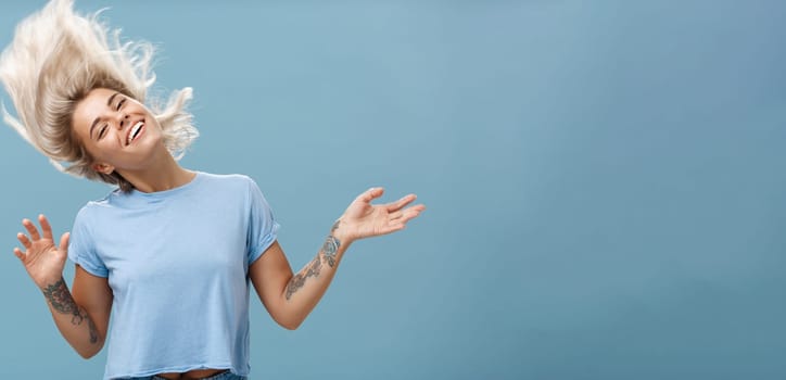 Waist-up shot of artistic and creative attractive carefree blonde female waving head and hair joyfully dancing and jumping having fun smiling broadly over blue background. Emotions and people concept