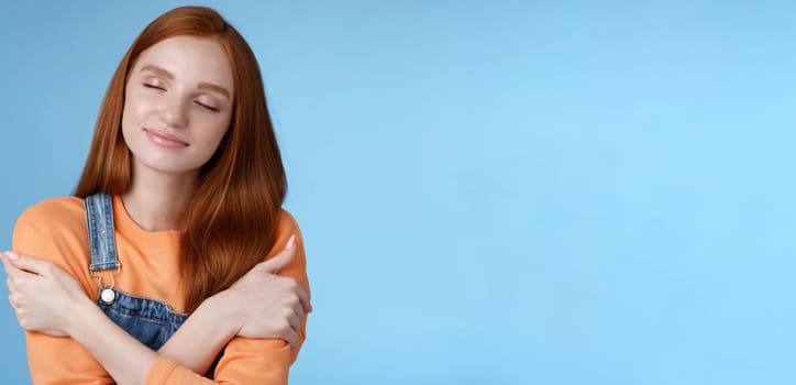 Passionate romantic tender ginger girl feel safe comfort close eyes smiling gently lovely daydreaming hugging herself recalling boyfriend cuddles sensual embraces, standing blue background happy.