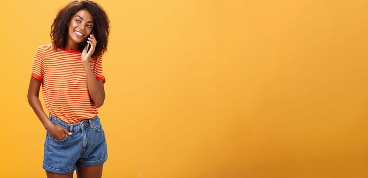 Stylish dark-skinned girl making casual phone call to friend telling all details of after romantic date standing pleased and carefree over orange background in striped t-shirt gazing left with grin. Lifestyle.