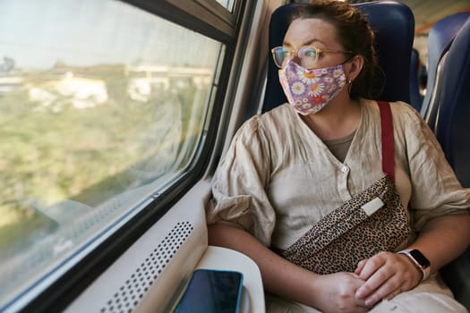 A girl in glasses and a medical mask riding a train and looking out the window. High quality photo