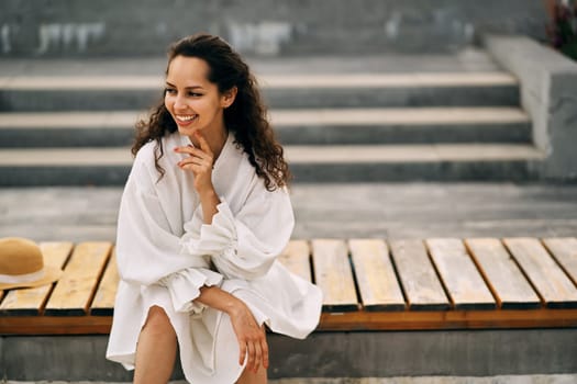 Smiling curly brunette girl in a white dress sitting on a wooden bench in the park. High quality photo