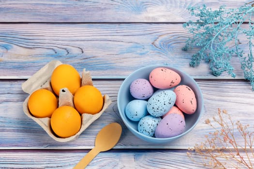 Bowl with colored Easter eggs and a cardboard box with fresh eggs, a wooden spoon on the boards with decorative plants. Top view.