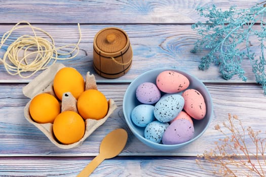 Bowl with colored Easter eggs and a cardboard box with fresh eggs, a small wooden barrel, a wooden spoon and a rope on the boards with decorative plants. Top view.