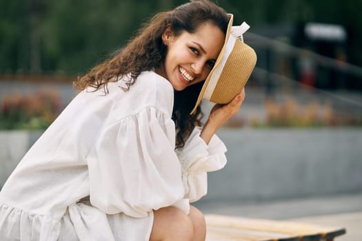 Smiling curly brunette girl in a white dress sitting and covering her face with a hat. High quality photo