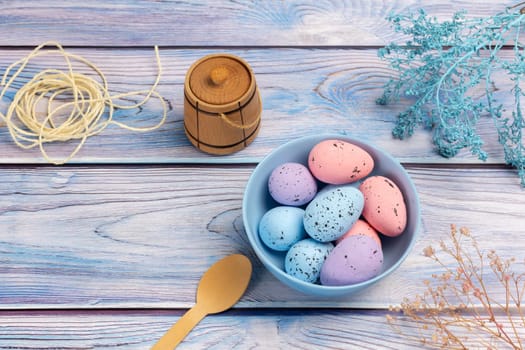 Bowl with colored Easter eggs, a small wooden barrel, a wooden spoon and a rope on the boards with decorative plants. Top view.