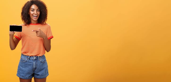 Girl showing picture in phone bragging about her cool vacation. Charming friendly and sociable african american woman with curly hair holding cellphone pointing at smartphone screen over orange wall. Technology and advertisement concept