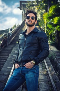 A man wearing a black jacket and sunglasses strikes a pose for a photo, exuding confidence and style. The background is simple, allowing the focus to remain on the subject.