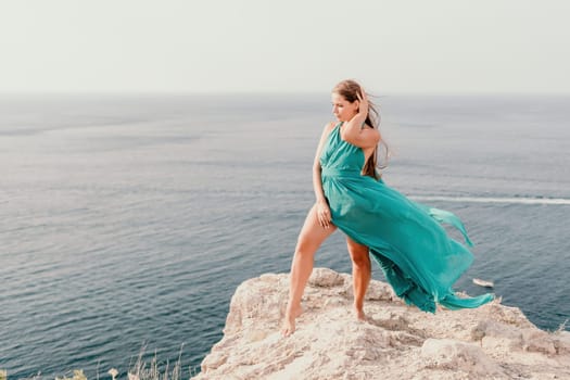 Side view a Young beautiful sensual woman in a mint long dress posing on a volcanic rock high above the sea during sunset. Girl on the nature on overcast sky background. Fashion photo