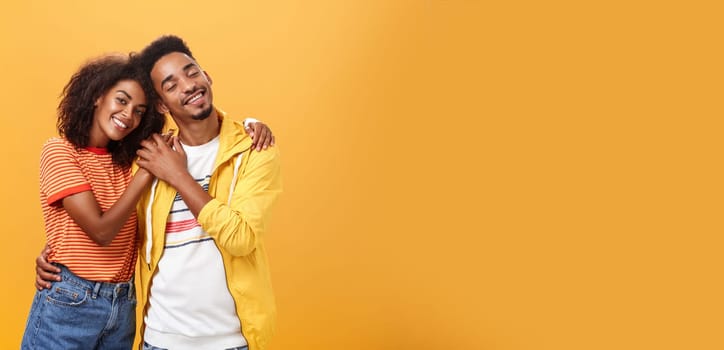 They love each other. Portrait of two charming african american man and woman in relationship hugging with heartwarming smile touching hands smiling gently posing against orange background.