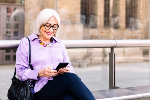 smiling senior woman sitting at the bus stop using the app on her mobile phone, concept of technology and elderly people leisure