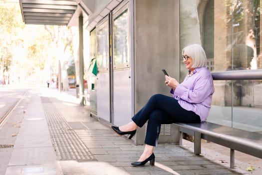 smiling senior woman waiting sitting at bus stop using app on mobile phone, concept of technology and elderly people leisure, copy space for text