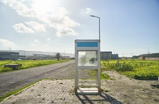 A phone booth stands alone on the grassy roadside, surrounded by a vast open field under a cloudy sky