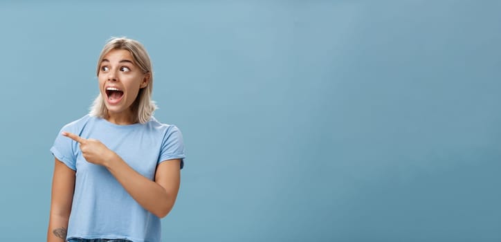 Girl being charmed and excited visiting awesome event pointing and staring left amazed and delighted standing happy and overwhelmed over blue background in trendy outfit dropping jaw. Emotions and advertising concept