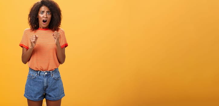 Woman about to panic hearing shocking worried news. Portrait of intense nervous and anxious dark-skinned adult girl with afro hairstyle clenching fists opening mouth looking concerned over orange wall.