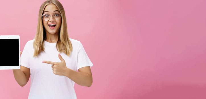 Girl suggests buy digital tablet for university and forget paper books. Excited happy and delighted young female student in white t-shirt and glasses pointing at device screen recommending gadget. Technology, emotions, advertising and lifestyle concept