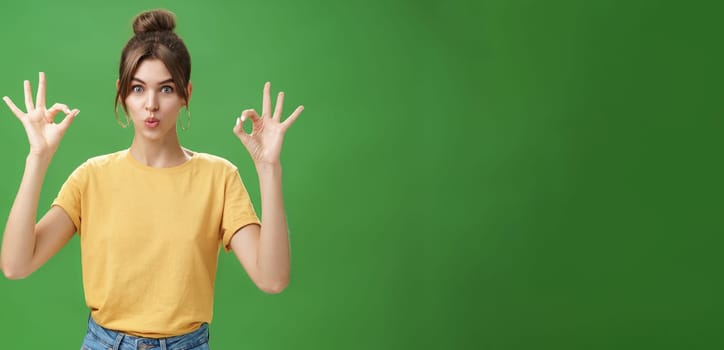 Portrait of silly cute and beautiful caucasian woman with combed hair in round earrings and yellow t-shirt showing okay, no problem gesture folding lips, reaffirming boss job goes well over green wall.