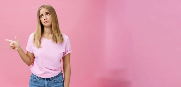 Envy upset cute young european woman with tanned skin and fair hait tilting head lifting eyebrows in sad silly look pointing, gazing left with regret and disappointment posing against pink background.