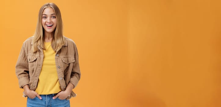 Charismatic tanned woman in corduroy jacket and yellow t-shirt ready for chilly autumn walk with friends smiling joyfully gazing entertained at camera holding hand in pockets casually over orange wall. Lifestyle.