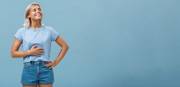 Cute girl likes eat after workout being pleased and stuffed after dinner. Portrait of satisfied attractive blonde young woman touching belly raising head up with broad smile over blue background.