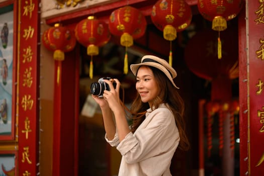 Smiling young woman standing at Chinese Temple with beautiful red lanterns adorning, taking photo with camera.