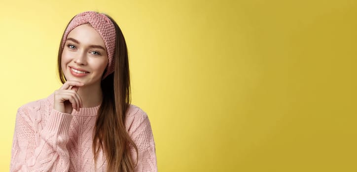 Happy young european woman in knitted headband, sweater tilting head touching chin and smiling silly, taking care of skin, feeling face glittering looking clear and healthy after skincare treatment.