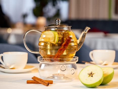 Herbal tea with apple and cinnamon in transparent glass teapot with warmer and candle on table in restaurant. Herbal tea concept, wellbeing and health care