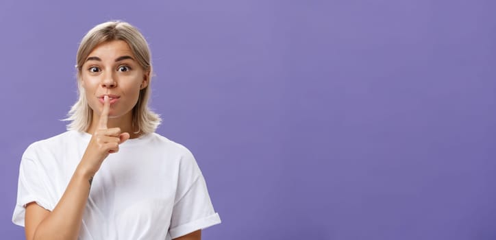 Lifestyle. Charming creative and stylish beautiful tanned girl with fair hair shushing at camera with delighted thrilled look making up great idea wanting hide it and keep secret posing over purple wall.