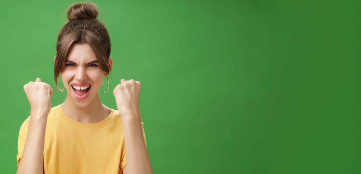 Woman with spirit of winner raising clenched fists smiling excited and supportive cheering being ready for gym excercised boosing confidents with yell looking daring at camera over green wall. Lifestyle.